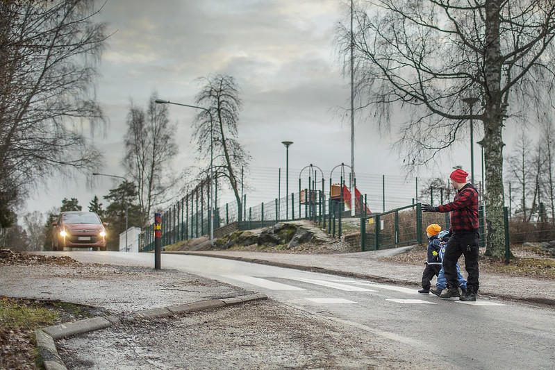 Perhe ylittämässä suojatietä leikkipuiston lähellä. Taustalla auto lähestymässä suojatietä.