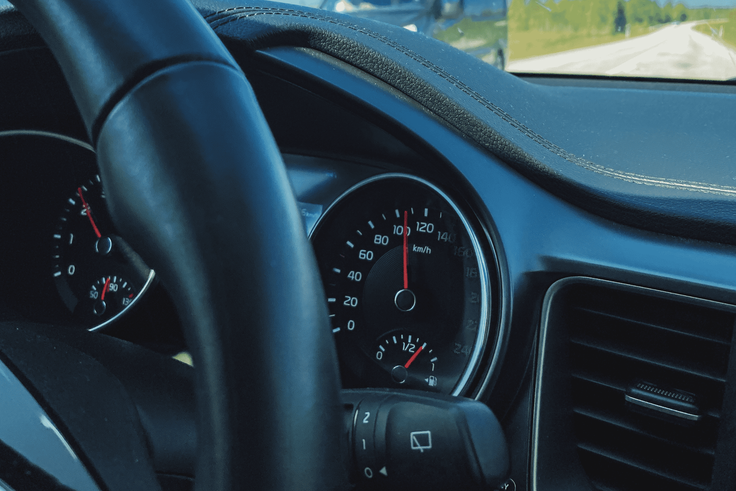 Auton mittaristo, joka näyttää ajonopeuden. Kuvan taustalla kesäinen maantie ja vastaantuleva auto.
